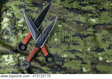 Black Throwing Knives On Camouflage Cloth. Three Special Knives With Red Handles Crossed. Military T