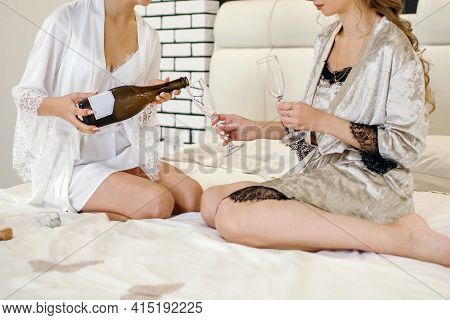 Two Girls In Beautiful Sexy Dressing Gowns Sit On The Bed And Drink Champagne. One Girl Is Pouring C