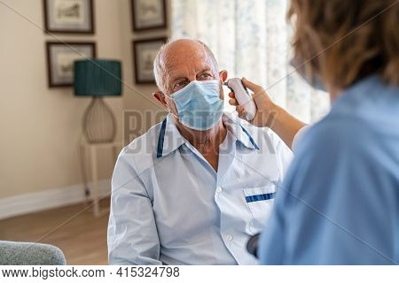 Senior man wearing face mask for safety against covid-19 and getting ear temperature checked by nurse at home. Nurse checking ear temperature of old patient using electronic tympanic thermometer. 
