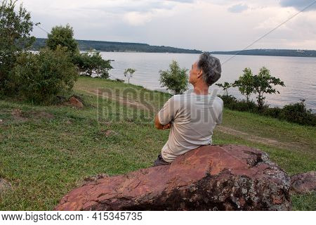 Senior Man At Parque Das Garças, In Brasilia, Leaning On A Big Rock While Looking Over Lake Paranoa