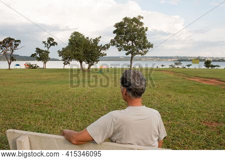Senior Man At Parque Das Garças, In Brasilia, Sitting On A Bench Enjoying Himself Looking Over Lake 