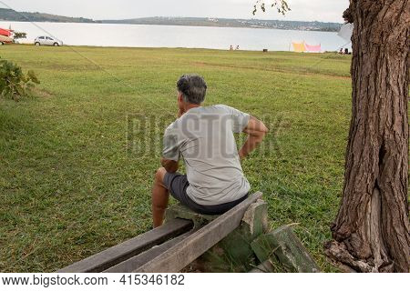 Senior Man At Parque Das Garças, In Brasilia, Sitting On A Bench Enjoying Himself Looking Over Lake 