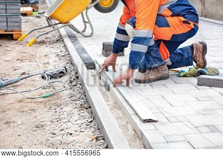 A Worker In A Blue-orange Overalls, Half-squatting, Measures Out A Part Of The Paving Slabs With A B