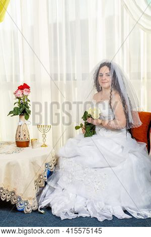 A Happy Jewish Bride With Her Face Covered With A Veil With A Bouquet Of White Roses Sits In A Synag