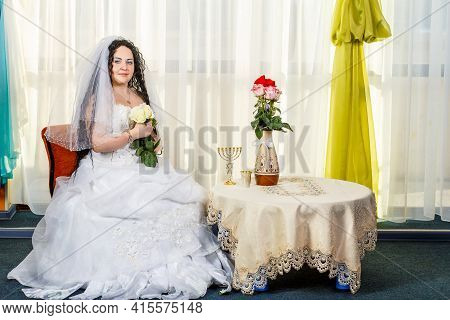 A Happy Jewish Bride With A Bouquet Of White Roses Sits In The Synagogue Before The Huppa Ceremony A
