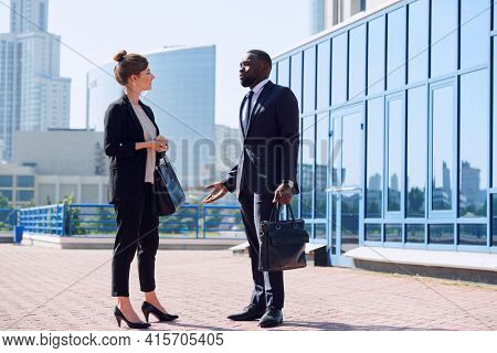 Two young intercultural businesspeople in suits discussing working moments against office building in urban environment on sunny day