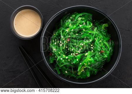 Healthy seaweed salad with sesame seeds in a black bowl and peanut sauce on the side on a black textured background. Top view.
