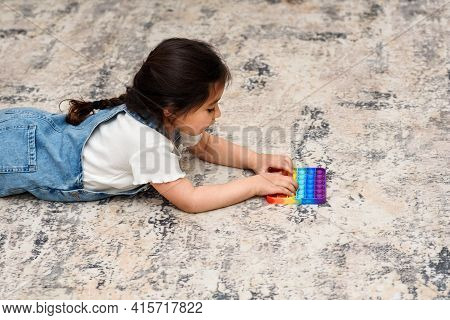 Young Little Girl Lies On The Floor At Home Playing Poppit-new Fidget Toy, Popular With Kids, Helps 