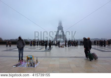 Paris, France - November 22, 2018: Eiffel Tower hidden in low clouds, Many people on Eiffel Tower viewing platform at Palais de Chaillot, Tourist with suitcase on wheels, Street souvenir vendor