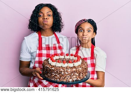 Beautiful african american mother and daughter wearing baker apron holding homemade cake puffing cheeks with funny face. mouth inflated with air, catching air. 