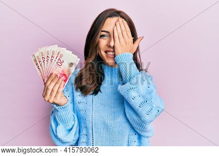 Young brunette woman holding 20 israel shekels banknotes covering one eye with hand, confident smile on face and surprise emotion. 