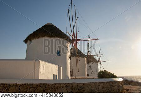 Old White Windmills And Stone Walls On The Coast Of Mykonos Island In Greece. In The Background The 