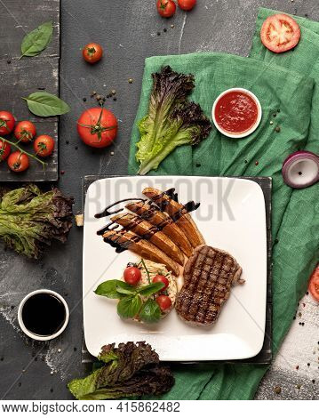 Grilled Meat Steak And Potatoes Cooking. White Square Food Plate On Kitchen Table Covered With Green