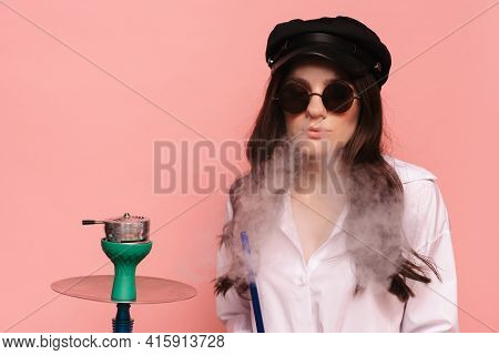 An Attractive Woman In A Cap And Glasses Smokes A Hookah, Sheesha And Enjoys Smoking. Pink Solid Col