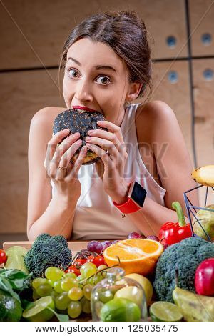 Hungry woman eating black burger at the table full of fruits and vegetables on the wooden background. Choosing between healthy and unhealthy food