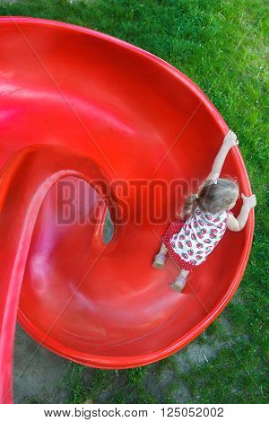 Overhead shot of little blonde girl is sliding down red plastic spiral playground slide