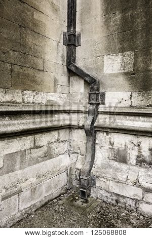 Architectural background of an old lead drainpipe winds its way down the corner of two stone walls to deliver water to the ground.