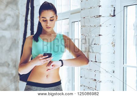 Checking all training updates. Beautiful young woman looking at her sport bracelet and using her smartphone while leaning to the wall at gym