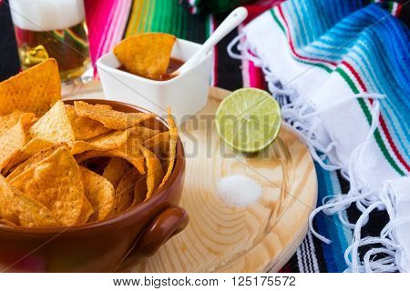 Nachos chips in a bowl over a chopping board with tomato sauce beer lime and salt with poncho background
