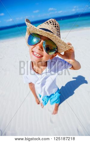 Happy boy in the sunglasses and hat on the tropical white beach