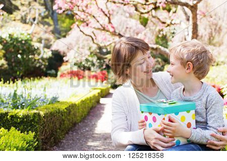 young beautiful mother and her adorable son sitting together in the park at spring time celebrating mother's day, boy giving her mother present and looking at each other