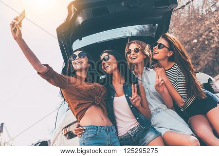 Just one selfie before road trip! Four beautiful young cheerful women making selfie with smile while standing near car trunk