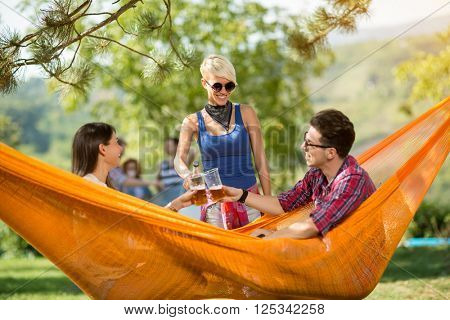 Young couple, male and female in hammock in nature toast with beer with female friend