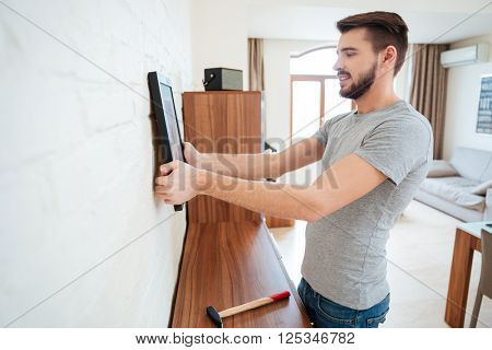 Concentrated handsome young man standing and hanging picture on white wall in the room