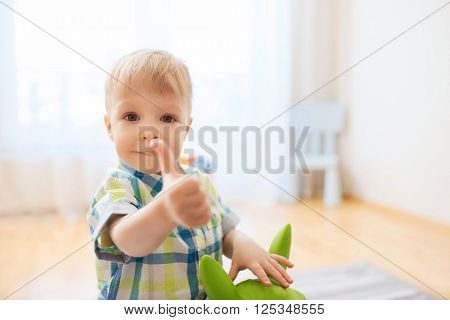 childhood, toys and people concept - happy little baby boy playing with ride-on toy horse and showing thumbs up at home