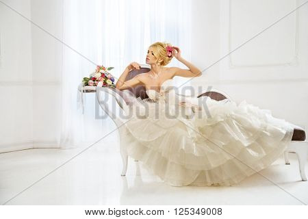 Beautiful fashion model. Sensual bride. Woman with wedding dress, hair and make up. Waiting for groom on background of window. Natural manicure. Beauty spring girl with bouquet of flowers in hands