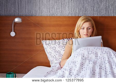 A young woman working on her tablet while lying in bed