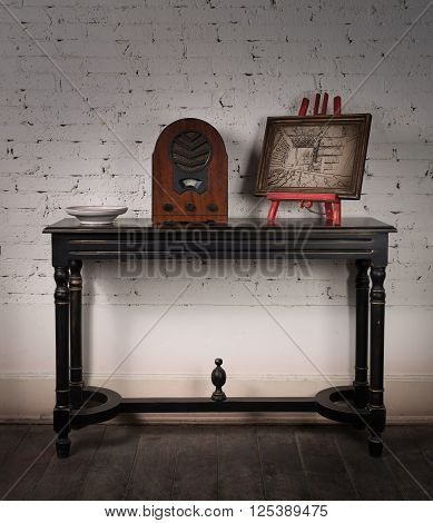 Vintage still life of an old analogue wooden radio and wooden framed painting on a black vintage wooden table and background of white painted brick wall and dark wooden floor