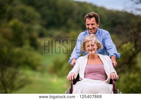 Senior man pushing woman sitting in wheelchair oustide in green autumn nature