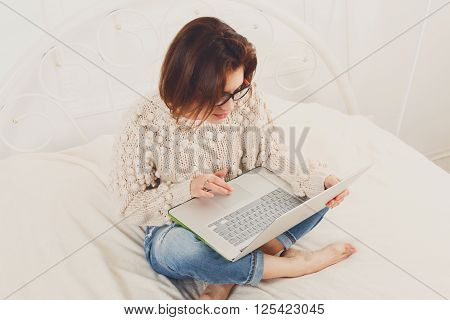 Young woman reads from laptop pc in bed. Engrossed Girl in eyeglasses looks into laptop. Beautiful woman in casual clothes at white bed surf internet at her computer. Freelancer. High key, soft tone