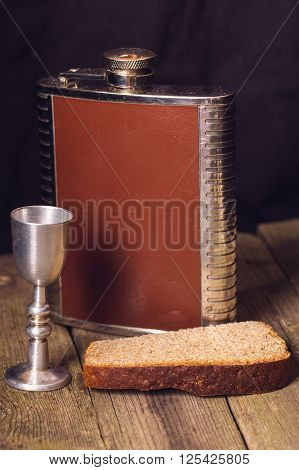 Flask shot glass and bread on a wooden table. The shot glass is turned from steel a table from old gray boards rye bread a dark background