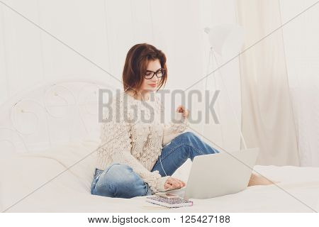 Young woman reads from laptop computer in bed. Engrossed Girl in eyeglasses and earphones with mobile phone. Attractive woman in casual clothes at white bed. Internet freelancer. High key, soft tone