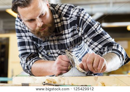 Hard working craftsman working with planer in a workshop for woodwork. Handsome man with tattoo and beard. Focus on the planer.