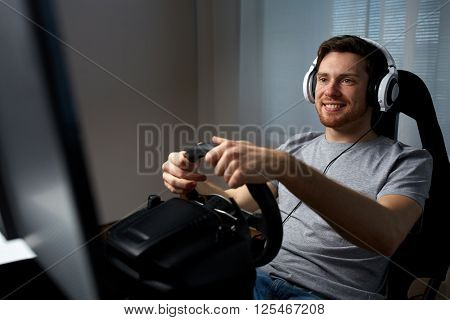 technology, gaming, entertainment and people concept - happy smiling young man in headphones with pc computer playing car racing video game at home and steering wheel