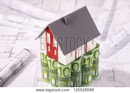 House stands on 100 euro banknotes surrounded by blueprints.