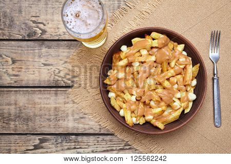 Poutine traditional Canadian meal with fries, curd cheese, beer and sauce on vintage wooden table background. Rustic style.