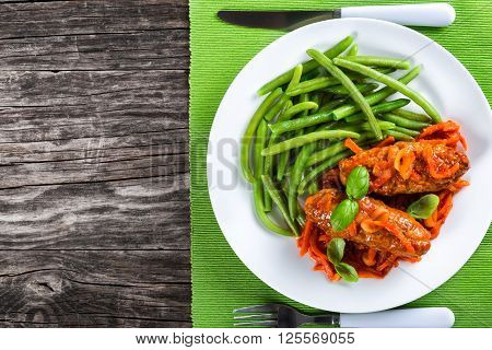Delicious Meat Cutlets stewed with onion carrot tomato sauce decorated with basil leaves in a white dish with boiled green bean on a table napkin with fork and knife top view