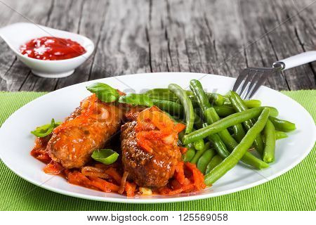 Delicious Meat Cutlets stewed with onion carrot tomato sauce decorated with basil leaves in a white dish with boiled green bean on a table napkin with fork top view