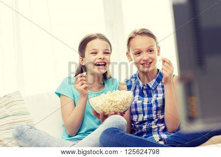 people, children, television, friends and friendship concept - two happy little girls watching comedy movie on tv and eating popcorn at home