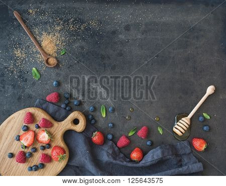 Berry frame with copy space on right. Strawberries, raspberries, blueberries and mint leaves, honey, cane sugar, dark grunge background, top view, copy space, horizontal