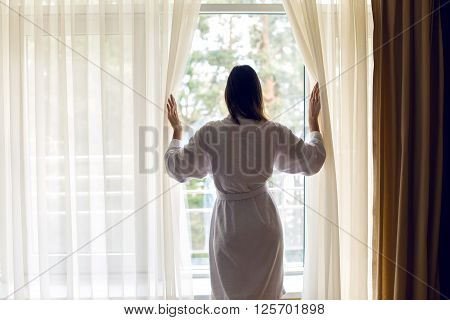 the girl standing at the window his back in a white Terry robe and opens the curtains