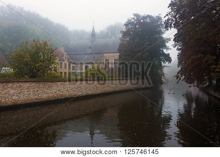 Begijnhof Bruges. Misty autumn morning haze over the water of canal and reflections of trees and ancient belgian houses in Minnerwater park in Brugge