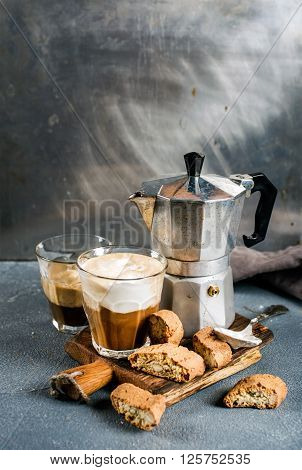 Glass of latte coffee on rustic wooden board, cantucci biscuits and steel Italian Moka pot, grey background, selective focus, copy space