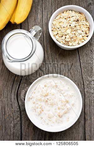 Oatmeal oats in bowl and jug of mik on wooden background top view