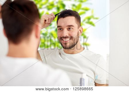 beauty, grooming and people concept - smiling young man looking to mirror and brushing hair with comb at home bathroom over green natural background