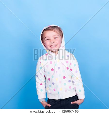 Laughing child girl 3-4 years old posing in room over blue. Wearing hoodie with polka dots. Hands in pockets. Looking at camera. Having fun.